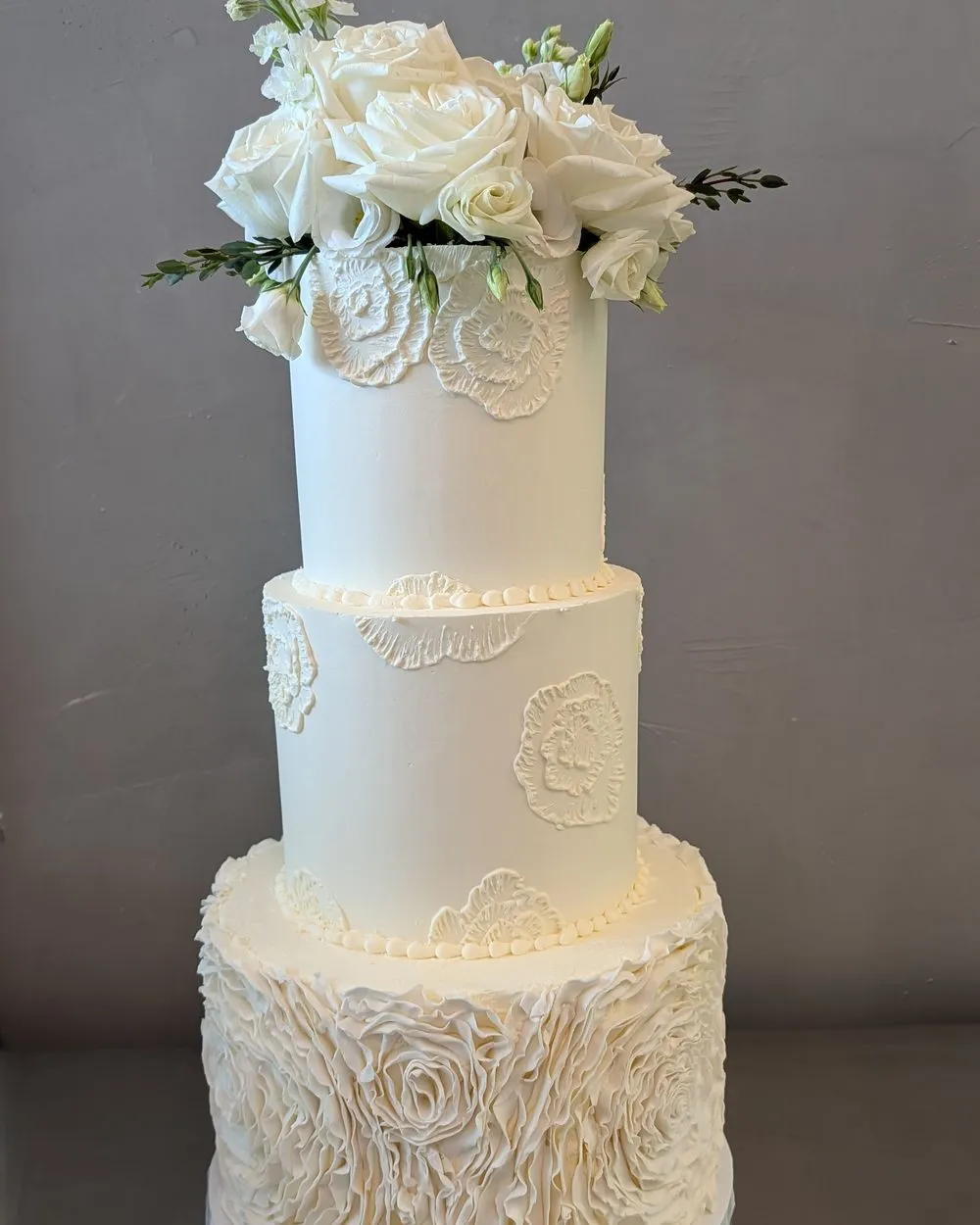 Three-tier ivory wedding cake with piped lace, a ruffled base tier and white roses